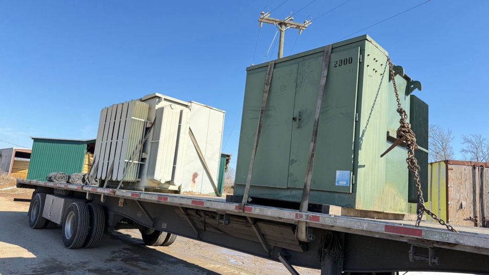 Two used green transformers chained and strapped down on a flatbed trailer, loaded out and ready to haul